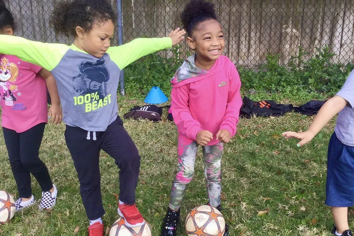 Children smiling and balancing on soccer balls during outdoor play in a fenced grassy area.